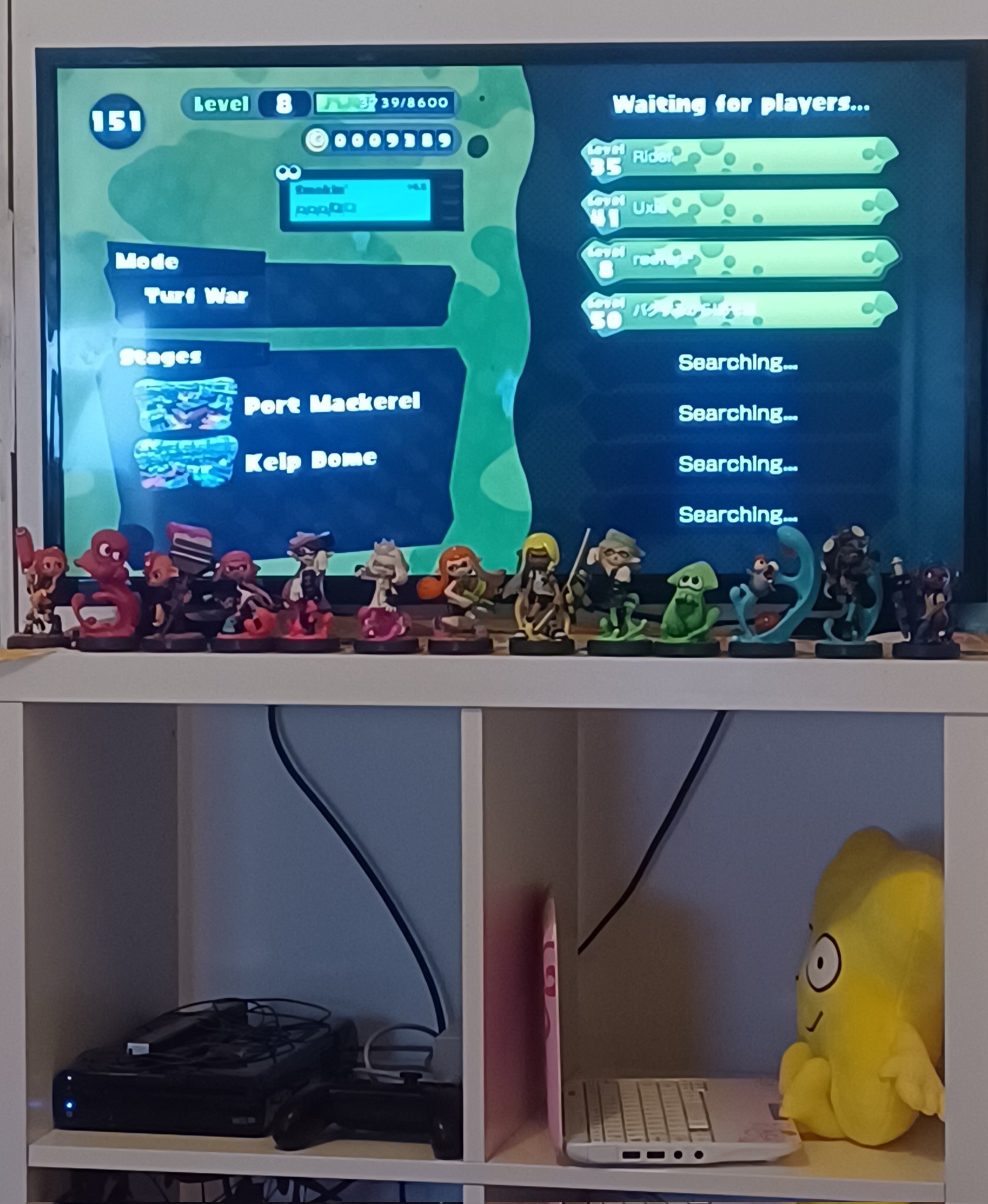 a photo of splatoon for wii u on a tv screen, in a Turf War lobby, with several splatoon amiibos in a rainbow line in front of it. the tv is on top of a stand which under it is separated into 4 compartments, although only the top 2 compartments are visible. in the leftmost compartment is the wii u, while in the rightmost compartment is a plushie of X from BFB at a disney asus EEEPC.